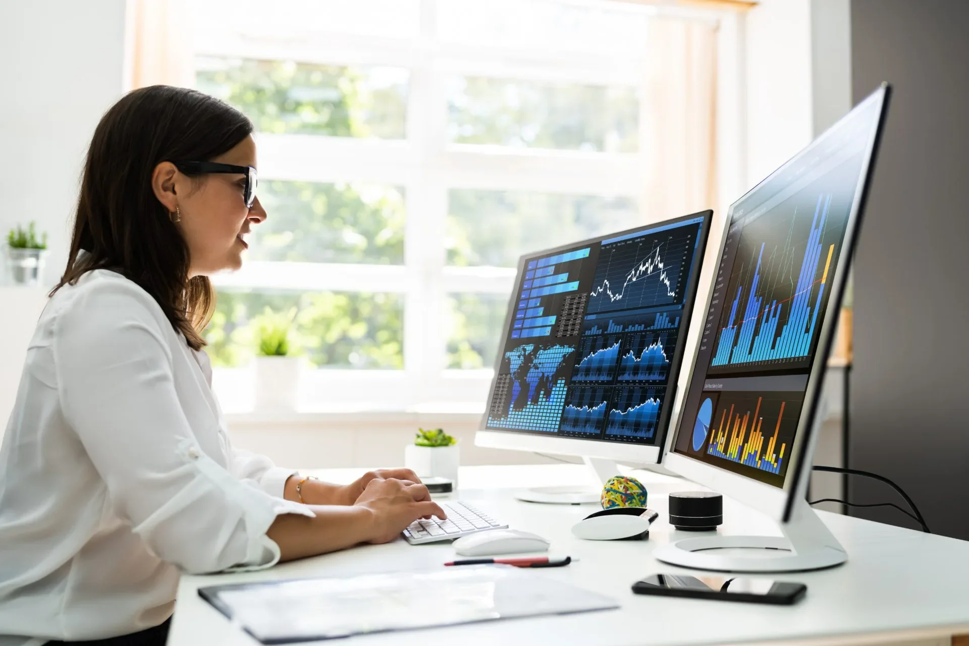 Woman at a desk looking at charts of data on two monitors.
